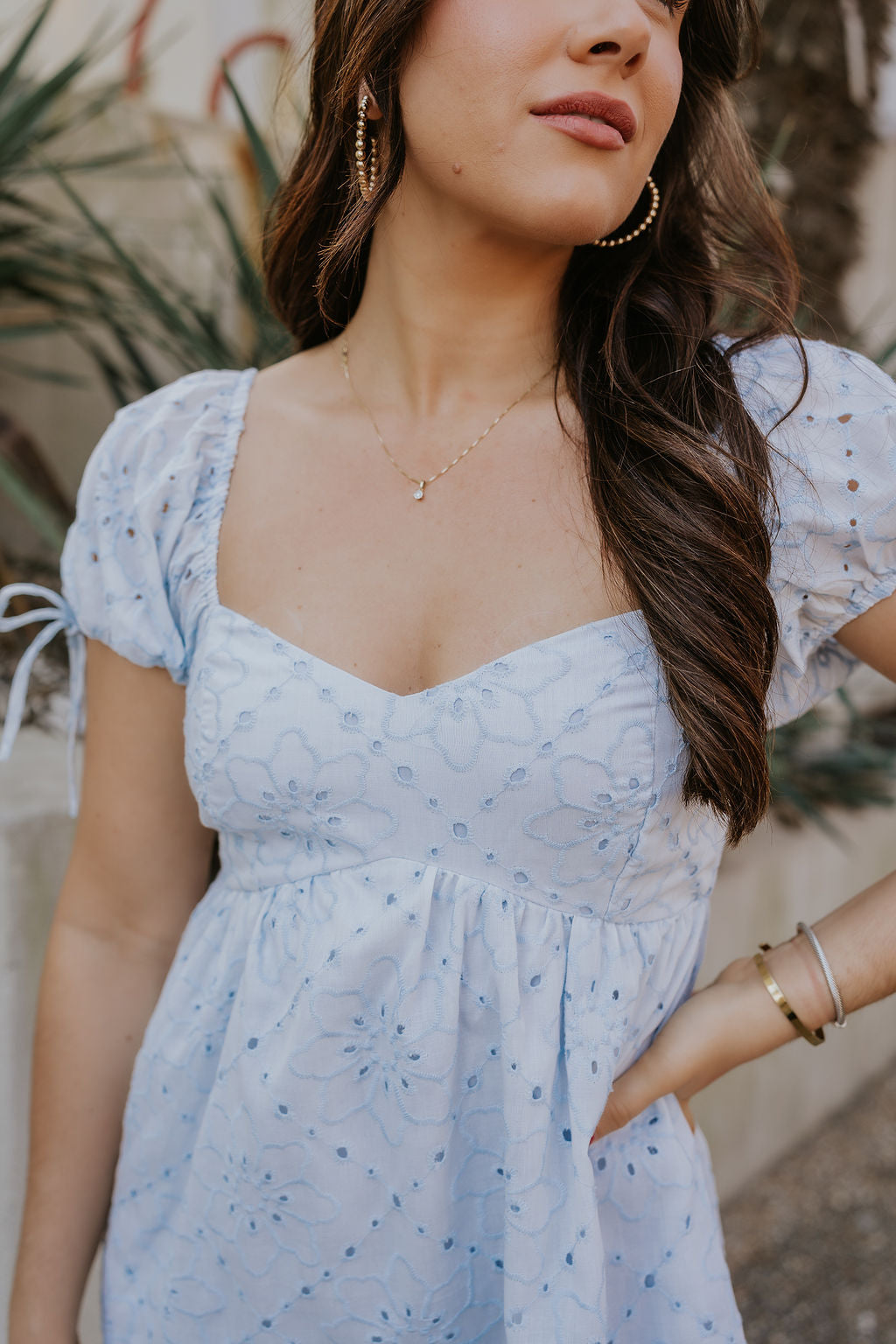 Close up view of female model wearing the The Ella Light Blue Eyelet Mini Dress which features light blue cotton fabric with an eyelet floral pattern, mini length, light blue lining, a sweetheart neckline, short puff sleeves with tie details, and a smocked back.