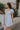 Side view of female model wearing the The Ella Light Blue Eyelet Mini Dress which features light blue cotton fabric with an eyelet floral pattern, mini length, light blue lining, a sweetheart neckline, short puff sleeves with tie details, and a smocked back.