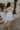 Full body back view of female model wearing the The Ella Light Blue Eyelet Mini Dress which features light blue cotton fabric with an eyelet floral pattern, mini length, light blue lining, a sweetheart neckline, short puff sleeves with tie details, and a smocked back.