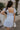 Back view of female model wearing the The Ella Light Blue Eyelet Mini Dress which features light blue cotton fabric with an eyelet floral pattern, mini length, light blue lining, a sweetheart neckline, short puff sleeves with tie details, and a smocked back.