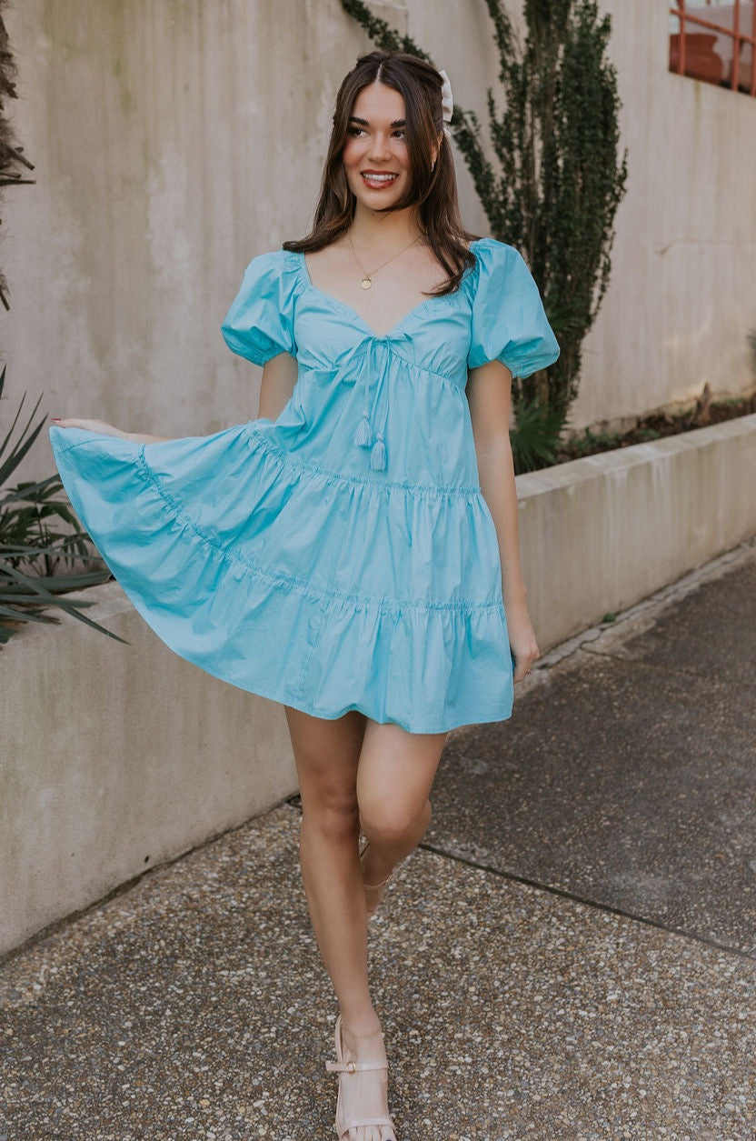 Full body view of female model wearing the Gloria Light Blue Short Sleeve Mini Dress which features aqua blue cotton fabric, aqua blue lining, mini length, ruffle tiered details, a sweetheart neckline with drawstring ties, a smocked back, and short puff sleeves.