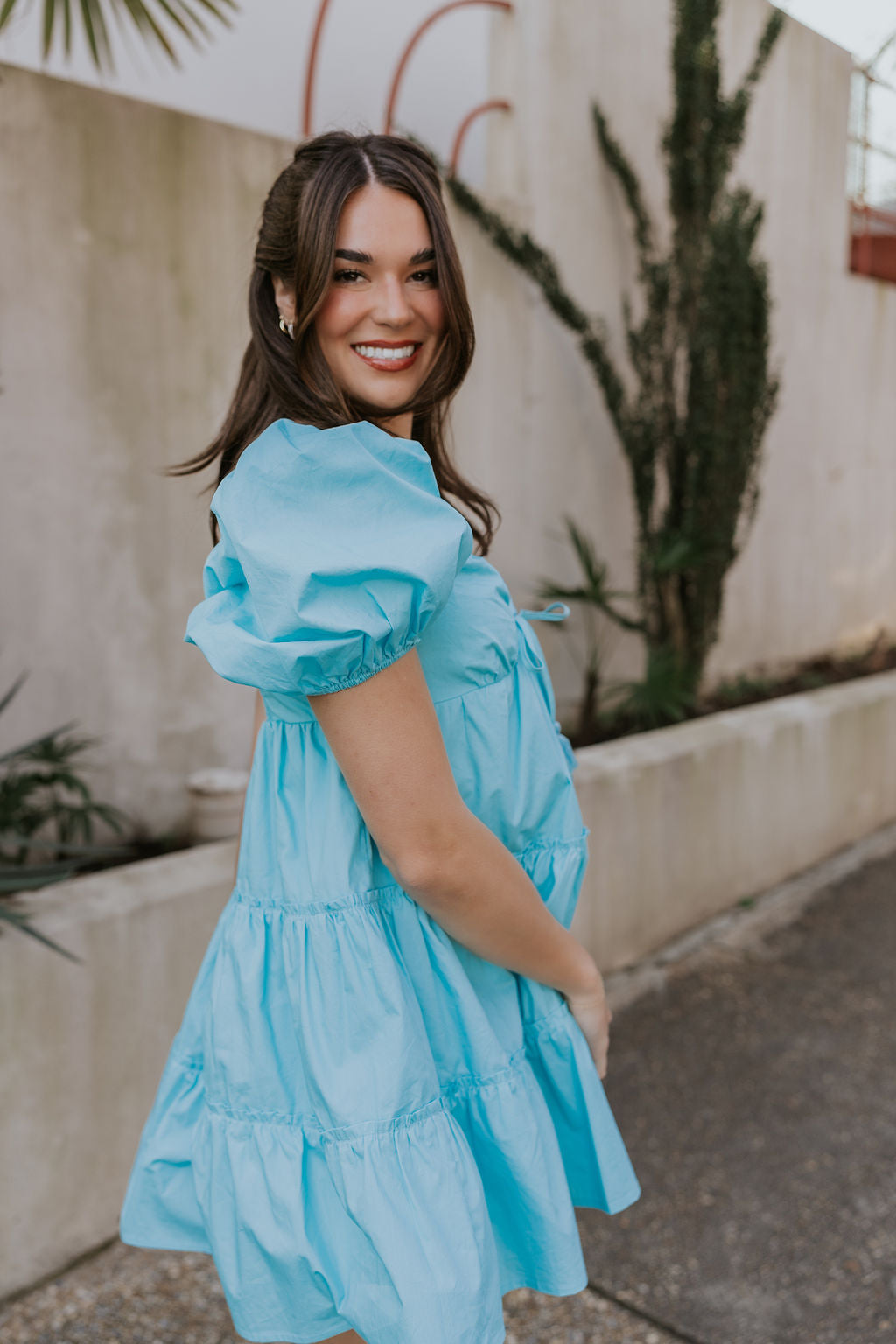 Side view of female model wearing the Gloria Light Blue Short Sleeve Mini Dress which features aqua blue cotton fabric, aqua blue lining, mini length, ruffle tiered details, a sweetheart neckline with drawstring ties, a smocked back, and short puff sleeves.