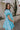 Side view of female model wearing the Gloria Light Blue Short Sleeve Mini Dress which features aqua blue cotton fabric, aqua blue lining, mini length, ruffle tiered details, a sweetheart neckline with drawstring ties, a smocked back, and short puff sleeves.