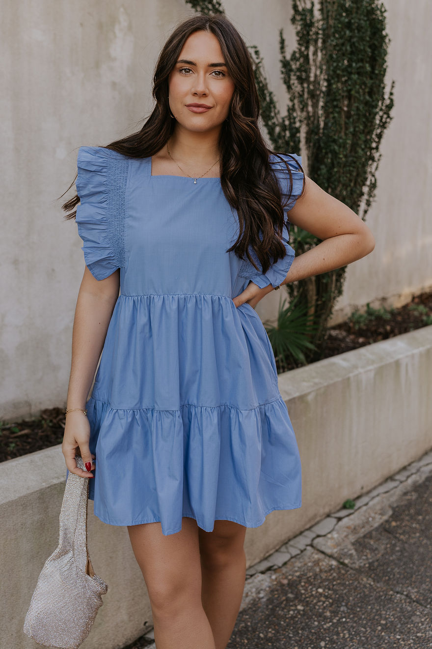 Front view of model wearing the Nicole Blue Ruffle Short Sleeve Mini Dress which features chambray blue light weight fabric, mini length, chambray blue lining, two-tiered design, two side pockets, square neckline and ruffle short sleeves.
