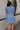 Back view of model wearing the Nicole Blue Ruffle Short Sleeve Mini Dress which features chambray blue light weight fabric, mini length, chambray blue lining, two-tiered design, two side pockets, square neckline and ruffle short sleeves.