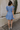 Full body back view of model wearing the Nicole Blue Ruffle Short Sleeve Mini Dress which features chambray blue light weight fabric, mini length, chambray blue lining, two-tiered design, two side pockets, square neckline and ruffle short sleeves.