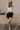 Full body view of model wearing the Tiffany Black Shorts which features black fabric with black lining, a front zipper with hook closures, side pockets, faux back pocket details, belt loops, and front pleats.