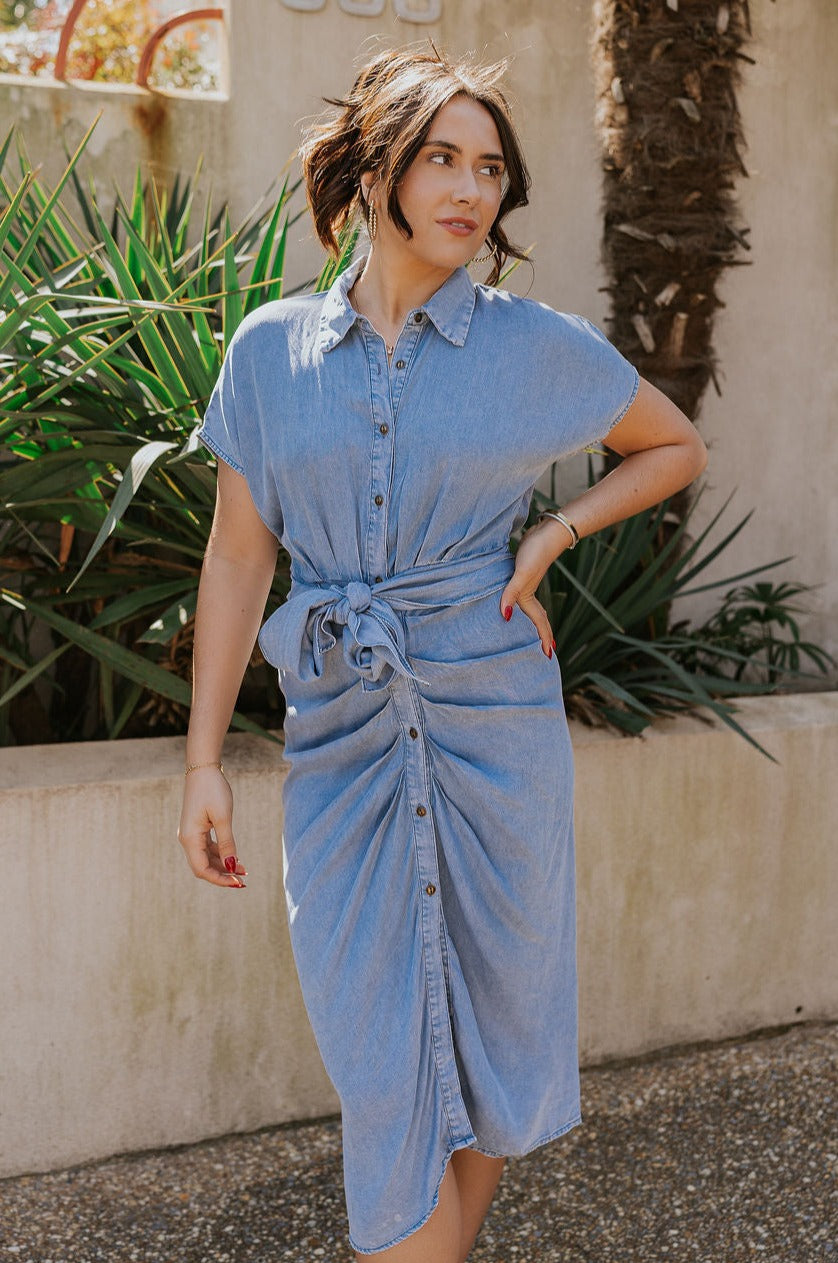 Front view of model wearing the Addison Washed Denim Front Tie Midi Dress which features washed denim fabric, midi length, brown tortoise button up front closure, front cinch tie waist, collared neckline and short sleeves.