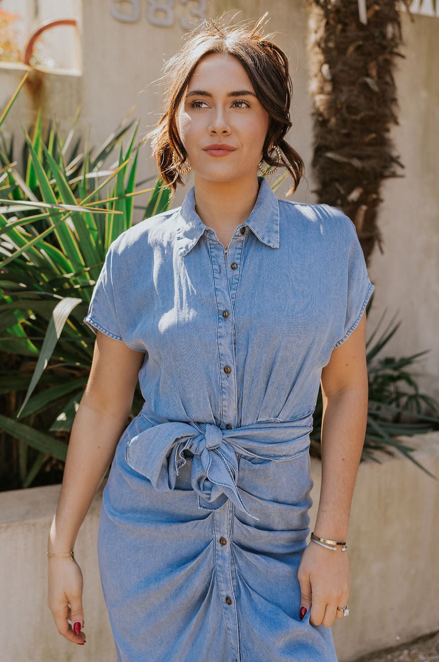 Front view of model wearing the Addison Washed Denim Front Tie Midi Dress which features washed denim fabric, midi length, brown tortoise button up front closure, front cinch tie waist, collared neckline and short sleeves.
