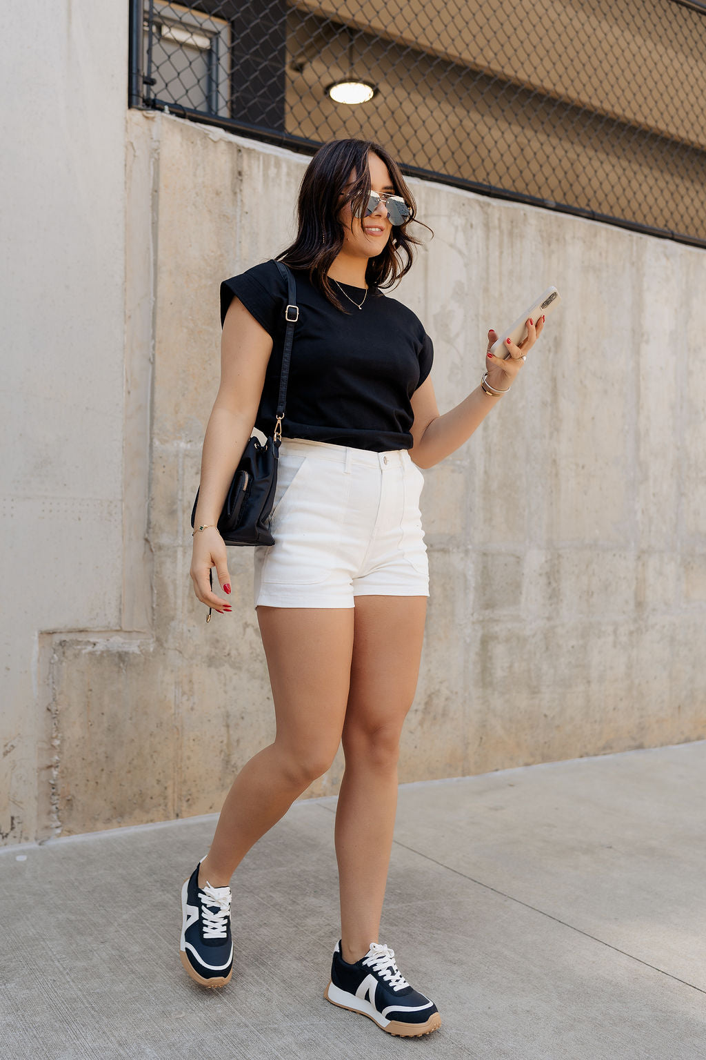 Full body view of model wearing the Iris Black Sleeveless Top that features black cotton fabric, a round neckline, and short sleeves with stitched ribbed hem details. Shirt is tucked into white shorts