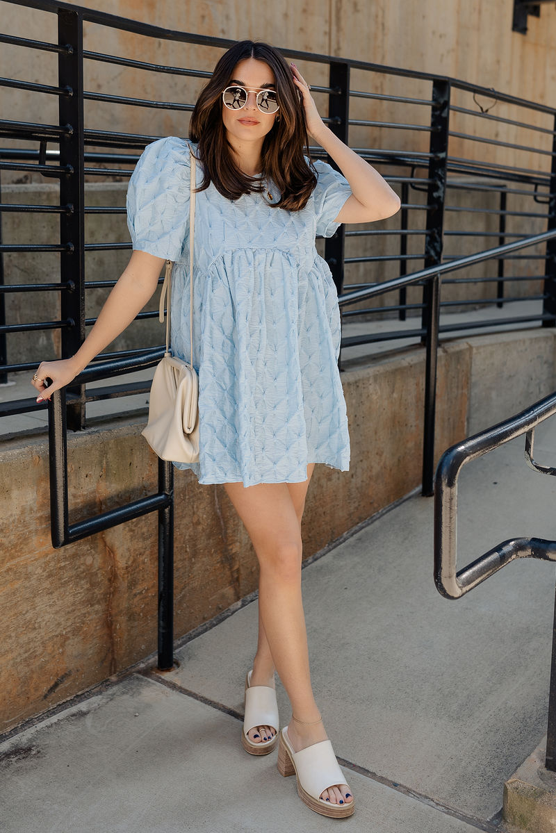 Full body view of model wearing the Amelia Light Blue Short Puff Sleeve BabyDoll Dress which features light blue textured fabric, bubble pattern design, mini length, round neckline, short puff sleeves and back button closure.
