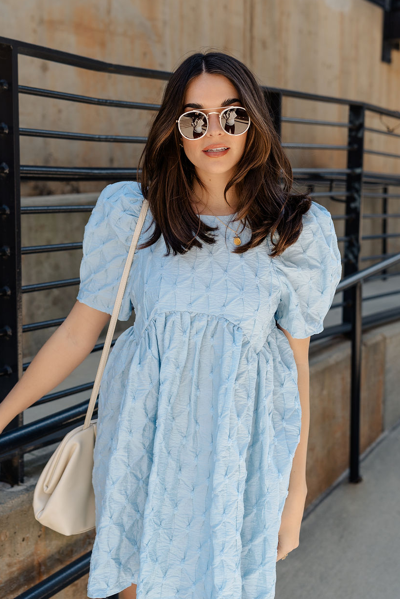 Front view of model wearing the Amelia Light Blue Short Puff Sleeve BabyDoll Dress which features light blue textured fabric, bubble pattern design, mini length, round neckline, short puff sleeves and back button closure.