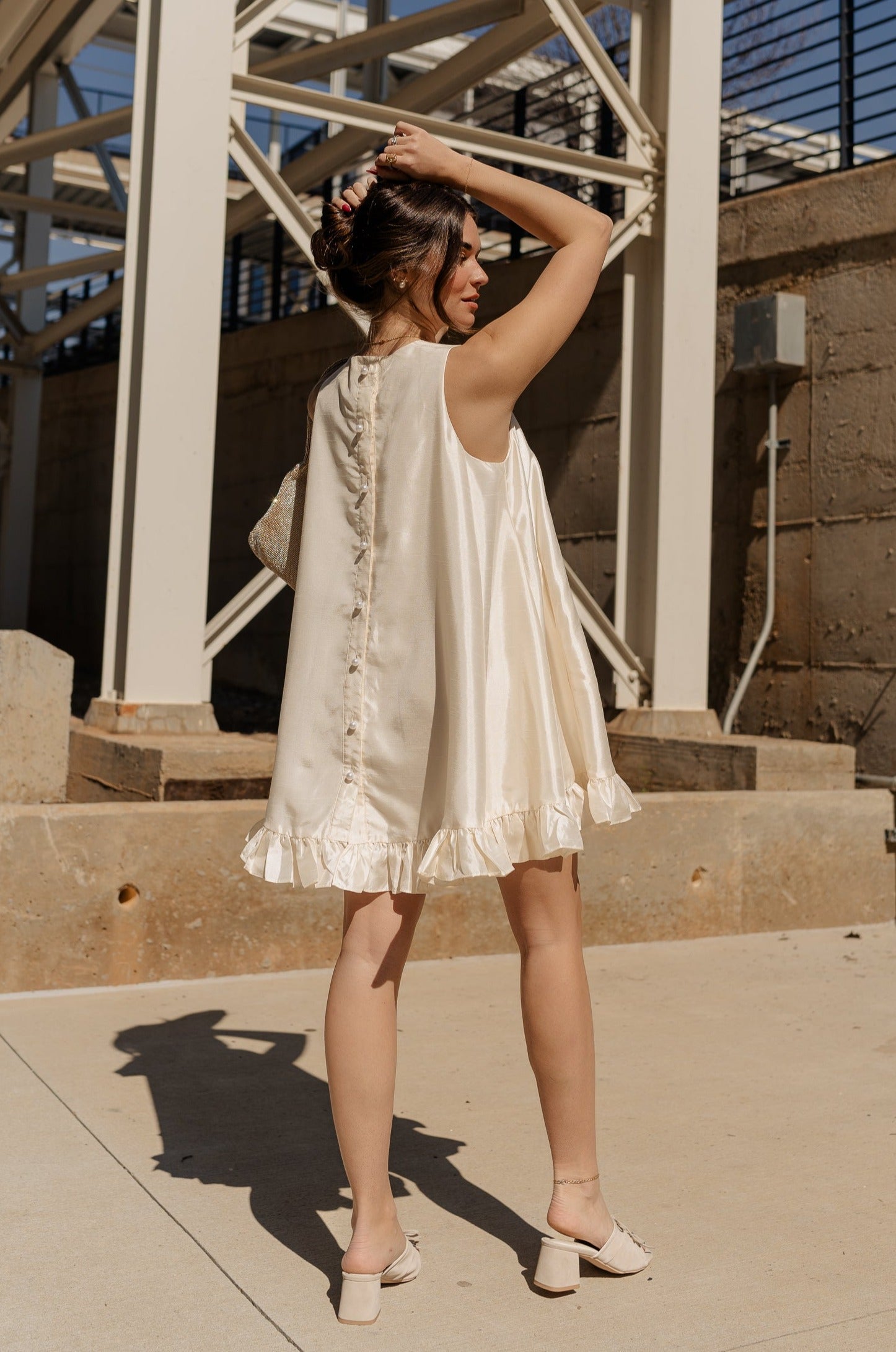 back image of model wearing the Anastasia Ruffle Mini Dress features cream sheen fabric, mini length, ruffle flare hem, cream lining, pockets on each side, round neckline, sleeveless and pearl button down closure on the back.
