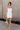 Full body front view of model wearing the Brielle White Sleeveless Mini Dress that features white stretchy fabric, mini length, white lining, a layered skirt, pleated cinched side detail, a square neckline, adjustable straps, and monochrome back zipper with a hook closure.