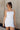 Front view of model wearing the Brielle White Sleeveless Mini Dress that features white stretchy fabric, mini length, white lining, a layered skirt, pleated cinched side detail, a square neckline, adjustable straps, and monochrome back zipper with a hook closure.
