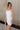 Side view of model wearing the Brielle White Sleeveless Mini Dress that features white stretchy fabric, mini length, white lining, a layered skirt, pleated cinched side detail, a square neckline, adjustable straps, and monochrome back zipper with a hook closure.