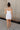 Full body back view of model wearing the Brielle White Sleeveless Mini Dress that features white stretchy fabric, mini length, white lining, a layered skirt, pleated cinched side detail, a square neckline, adjustable straps, and monochrome back zipper with a hook closure.