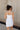 Back view of model wearing the Brielle White Sleeveless Mini Dress that features white stretchy fabric, mini length, white lining, a layered skirt, pleated cinched side detail, a square neckline, adjustable straps, and monochrome back zipper with a hook closure.