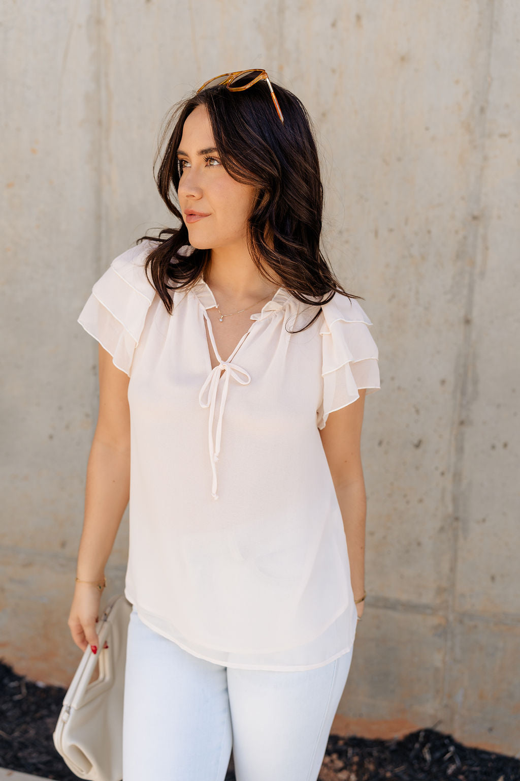 Frontal side view of model wearing the Olivia Cream Ruffle Short Sleeve Top which features cream sheer fabic, cream lining, ruffle neckline with drawstring ties, and ruffle tiered short sleeves.