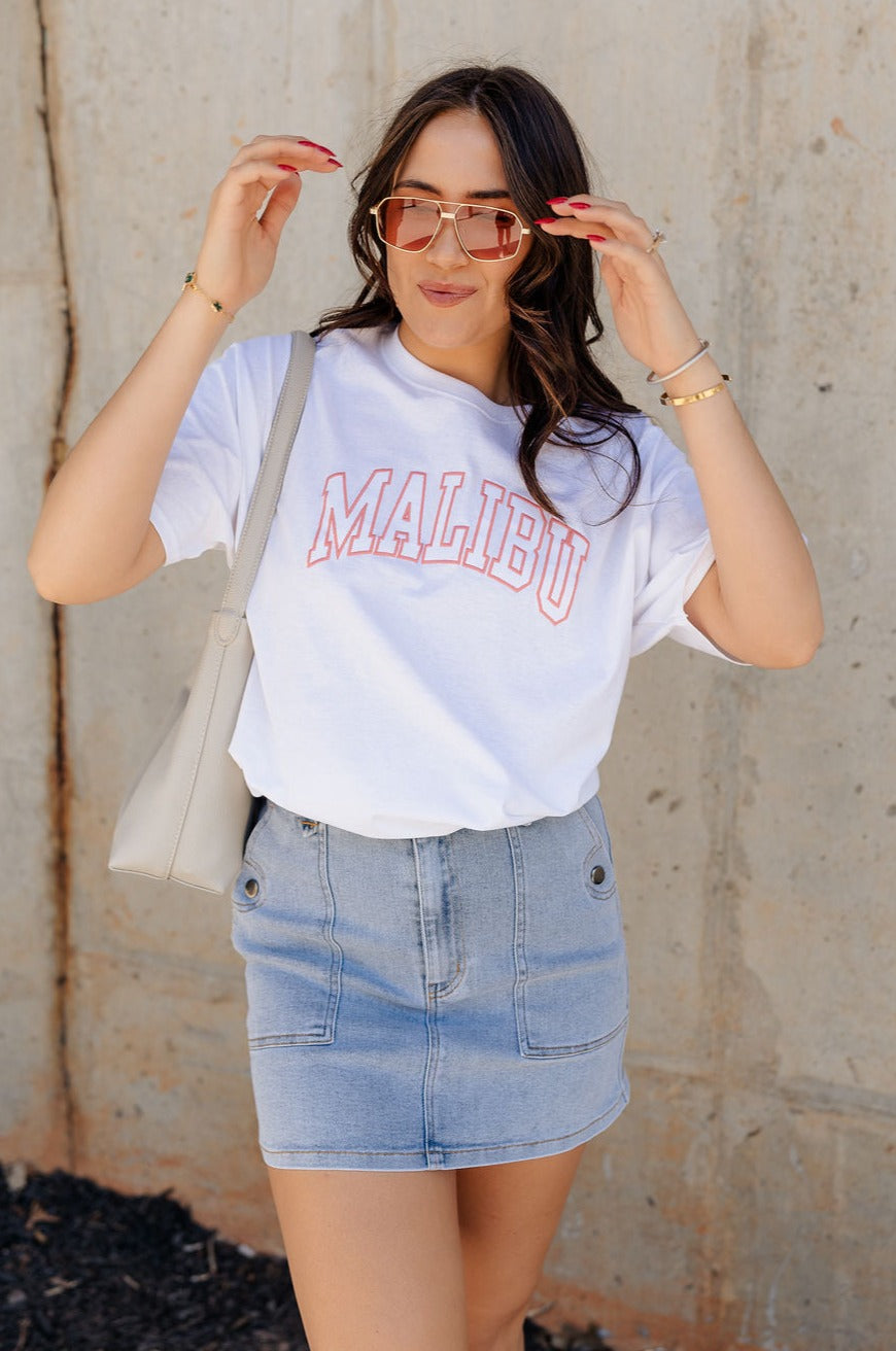 Front view of model wearing the Malibu White & Blush Short Sleeve Graphic Top which features white knit fabic, round neckline, shirt sleeves. Graphic says "MALIBU" in block letters with blush pink stitch.