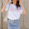 Front view of model wearing the Malibu White & Blush Short Sleeve Graphic Top which features white knit fabic, round neckline, shirt sleeves. Graphic says "MALIBU" in block letters with blush pink stitch.