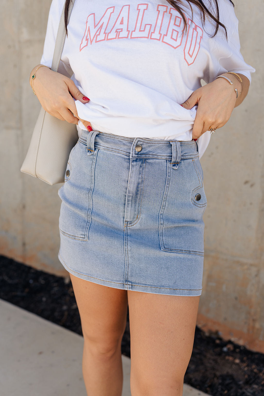 Front view of model wearing the Sloane Light Denim Wash Skirt which features light wash stretchy denim fabric, a front zipper with a snap closure, large side pockets with snap closures, back pockets, belt loops, and contrast stitching.