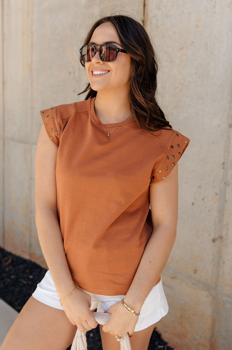 Front view of model wearing the Rylee Rust Sleeveless Top which features rust brown knit fabric, round neckline and sleeveless with gold stud details.