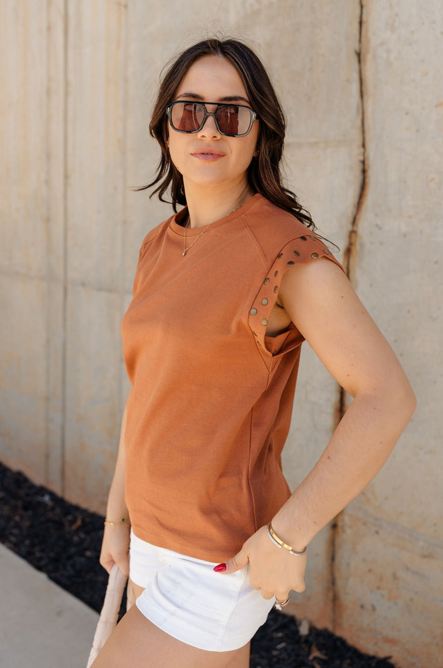 Side view of model wearing the Rylee Rust Sleeveless Top which features rust brown knit fabric, round neckline and sleeveless with gold stud details.