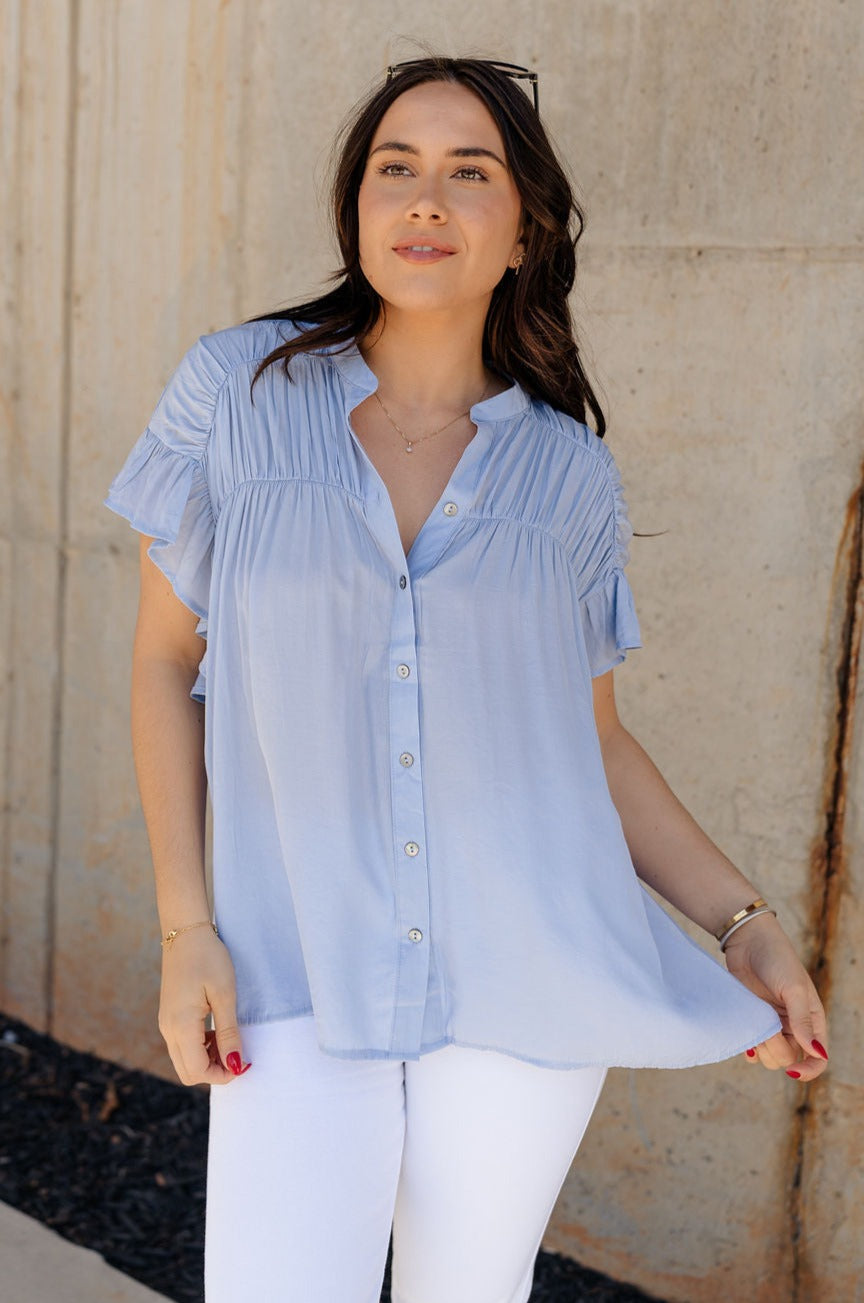 Front view of model wearing the Kennedy Light Blue Button-Up Top which features light blue fabric, monochrome button up front closure, round neckline with v-neckline cutout and short ruffle sleeves.