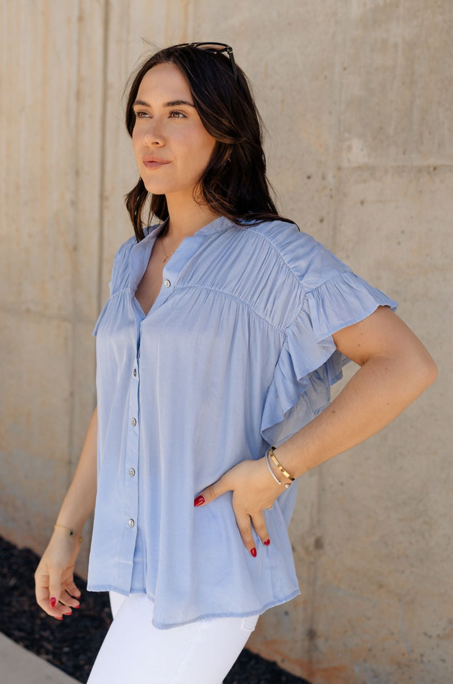 Side view of model wearing the Kennedy Light Blue Button-Up Top which features light blue fabric, monochrome button up front closure, round neckline with v-neckline cutout and short ruffle sleeves.