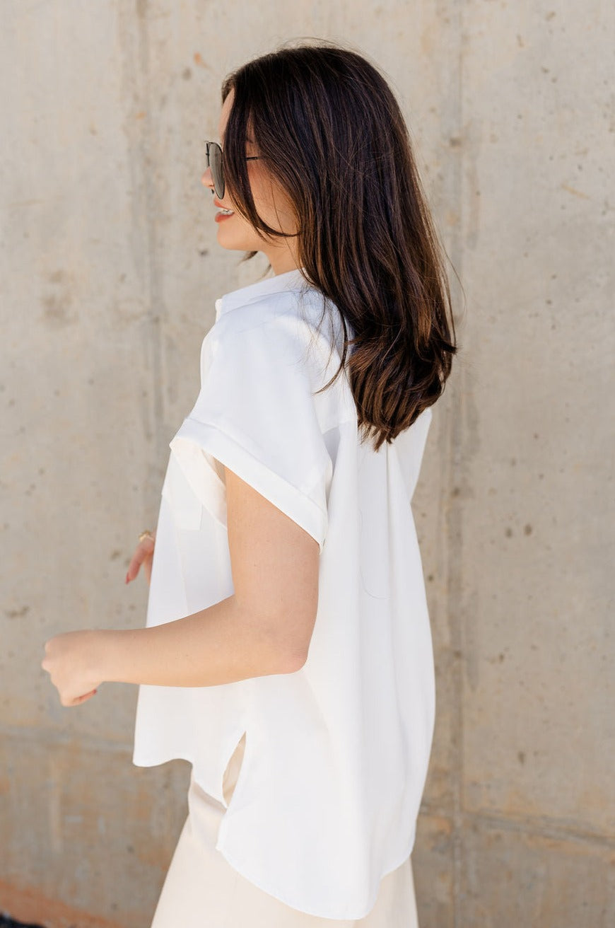 Side view of model wearing the Claire Off White Collared Short Sleeve Top which features white fabric, a front chest pocket, short sleeves with cuffs, small side slits, a button-up front, and a collared neckline.