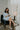 Model is sitting and reading while wearing the semi-sheer ivory fabric with subtle plaid texture, an abstract blue and green print, shimmer thread details, a surplice neckline, an elastic waist, a layered skirt, un-lined 3/4 sleeves with button closures, and ivory lining in the top and skirt.