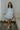 Full body front view of model wearing the semi-sheer ivory fabric with subtle plaid texture, an abstract blue and green print, shimmer thread details, a surplice neckline, an elastic waist, a layered skirt, un-lined 3/4 sleeves with button closures, and ivory lining in the top and skirt.