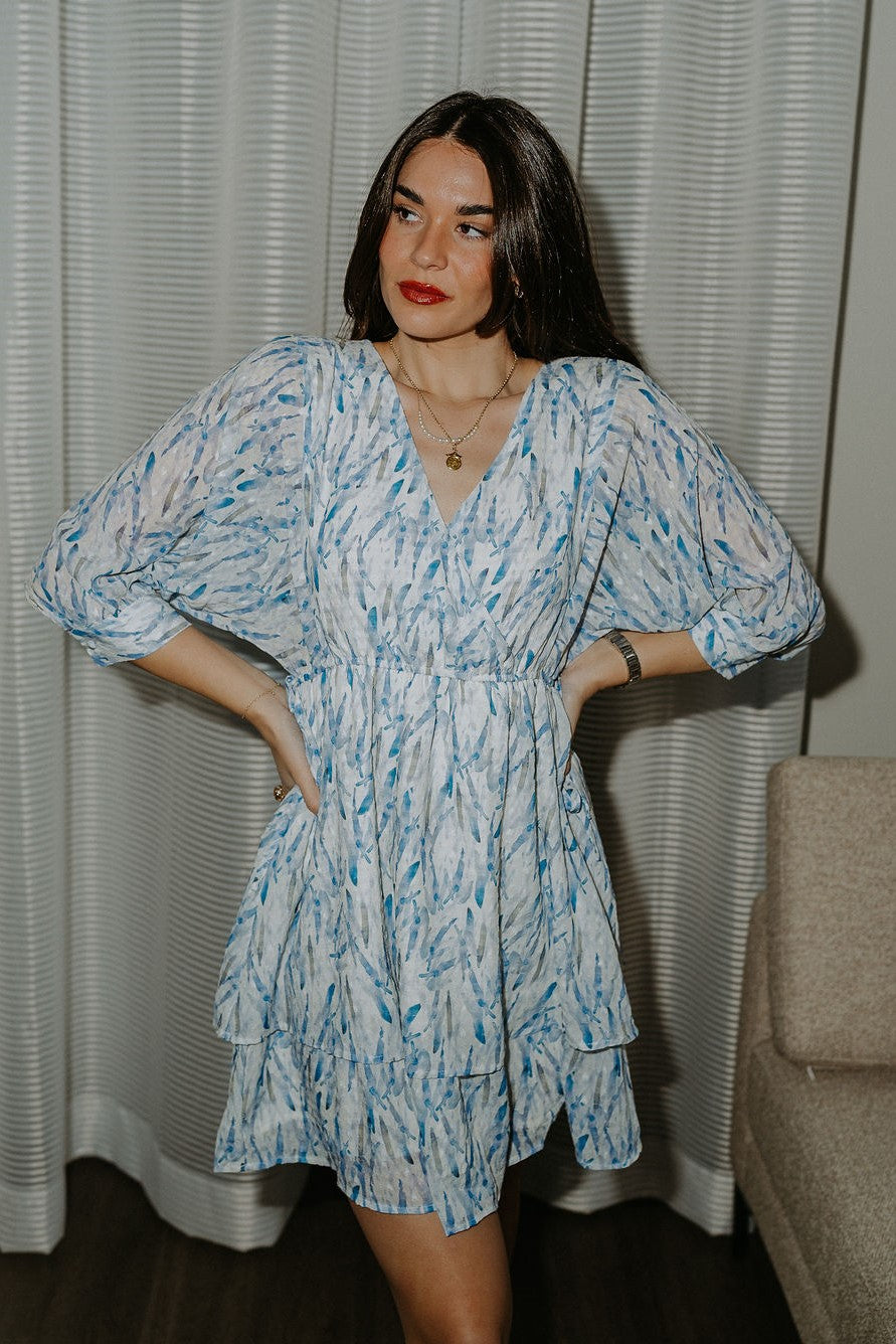 Front view of model wearing the Aurora Ivory & Blue Printed Mini Dress that  features semi-sheer ivory fabric with subtle plaid texture, an abstract blue and green print, shimmer thread details, a surplice neckline, an elastic waist, a layered skirt, un-lined 3/4 sleeves with button closures, and ivory lining in the top and skirt.