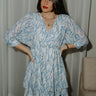 Front view of model wearing the Aurora Ivory & Blue Printed Mini Dress that  features semi-sheer ivory fabric with subtle plaid texture, an abstract blue and green print, shimmer thread details, a surplice neckline, an elastic waist, a layered skirt, un-lined 3/4 sleeves with button closures, and ivory lining in the top and skirt.