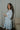 Side view of model wearing the semi-sheer ivory fabric with subtle plaid texture, an abstract blue and green print, shimmer thread details, a surplice neckline, an elastic waist, a layered skirt, un-lined 3/4 sleeves with button closures, and ivory lining in the top and skirt.