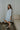 Full body side view of model wearing the semi-sheer ivory fabric with subtle plaid texture, an abstract blue and green print, shimmer thread details, a surplice neckline, an elastic waist, a layered skirt, un-lined 3/4 sleeves with button closures, and ivory lining in the top and skirt.