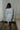 Full body back view of model wearing the semi-sheer ivory fabric with subtle plaid texture, an abstract blue and green print, shimmer thread details, a surplice neckline, an elastic waist, a layered skirt, un-lined 3/4 sleeves with button closures, and ivory lining in the top and skirt.