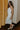 Full body side view of model wearing the Camila Cream Denim Button-Up Midi Dress which features  cream denim fabric, contrast stitching, a button-up front with silver button closures, side and back pockets, a sweetheart neckline, thick straps, and a midi-length hem.