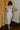 Full body back view of model wearing the Camila Cream Denim Button-Up Midi Dress which features  cream denim fabric, contrast stitching, a button-up front with silver button closures, side and back pockets, a sweetheart neckline, thick straps, and a midi-length hem.