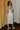 Full body back view of model wearing the Camila Cream Denim Button-Up Midi Dress which features  cream denim fabric, contrast stitching, a button-up front with silver button closures, side and back pockets, a sweetheart neckline, thick straps, and a midi-length hem.