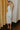 Full body back view of model wearing the Elisya Cream Ruched Midi Dress which features cream sheen knit fabric, midi length, cream lining, slit on the side, ruched side details, high neckline and sleeveless.
