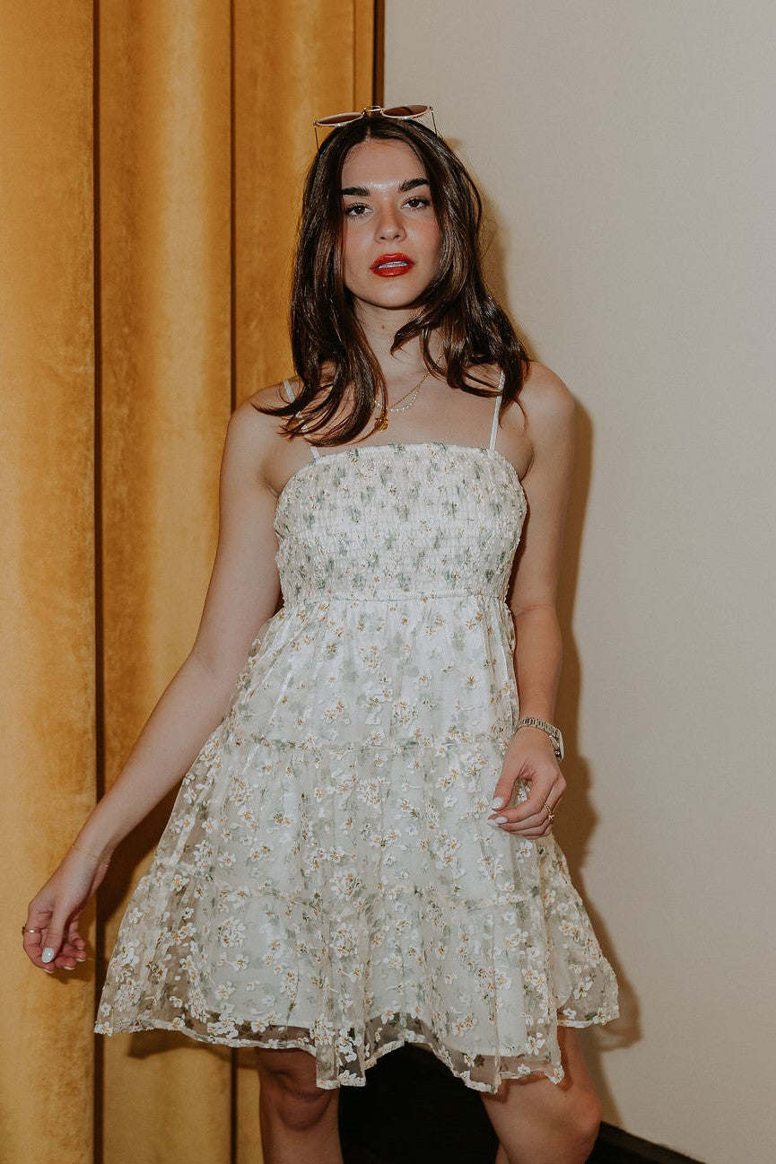 front view of model wearing the Angelica Cream Floral Mini Dress that features cream mesh fabric with a light green leaf pattern and embroidered white and gold flowers, cream lining, a smocked top, and adjustable spaghetti straps.
