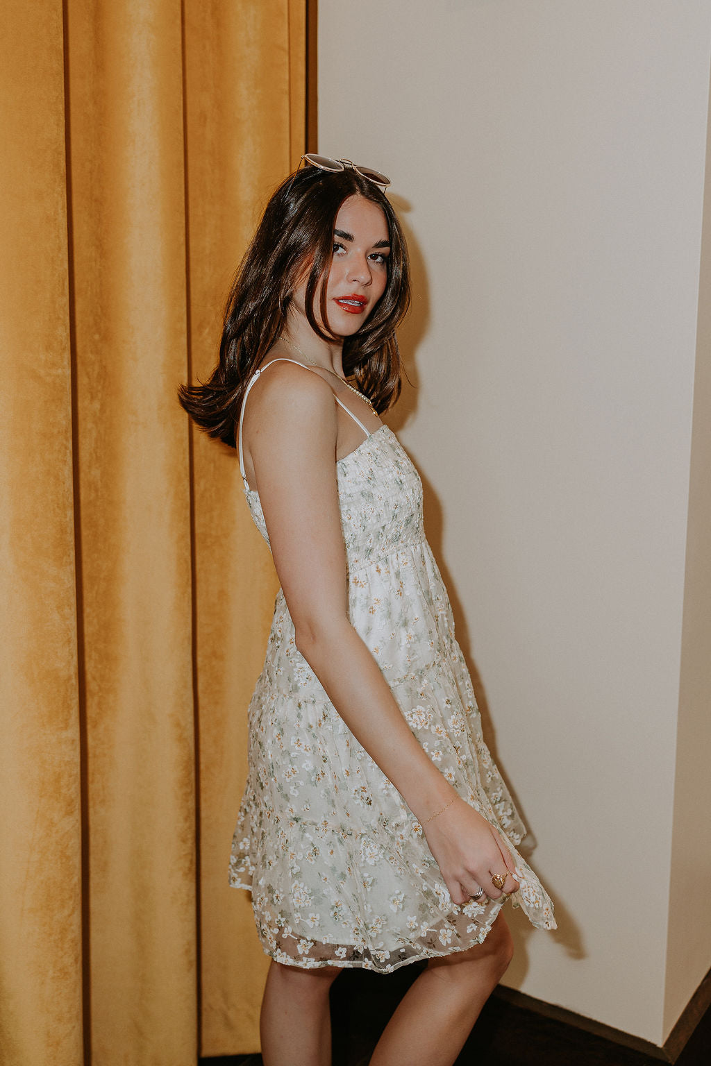 Side view of model wearing the Angelica Cream Floral Mini Dress that features cream mesh fabric with a light green leaf pattern and embroidered white and gold flowers, cream lining, a smocked top, and adjustable spaghetti straps.