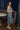 Full body side view of model wearing the Lucia Acid Indigo Denim Midi Dress which features acid medium wash denim fabric, midi length, silver button-up front closure, slit in the back, two back pockets, belt loops, sweetheart neckline, straps and open back.