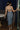 Full body back view of model wearing the Lucia Acid Indigo Denim Midi Dress which features acid medium wash denim fabric, midi length, silver button-up front closure, slit in the back, two back pockets, belt loops, sweetheart neckline, straps and open back.