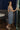 Full body back view of model wearing the Lucia Acid Indigo Denim Midi Dress which features acid medium wash denim fabric, midi length, silver button-up front closure, slit in the back, two back pockets, belt loops, sweetheart neckline, straps and open back.