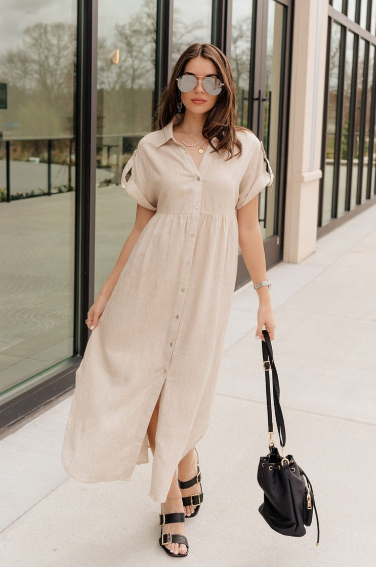 Full body view of model wearing the Daphne Oatmeal Button-Up Midi Dress which features beige fabric, a button-up front with pearlescent buttons, a collared neckline, short sleeves with cuffs and button tabs, and a rounded midi hemline.