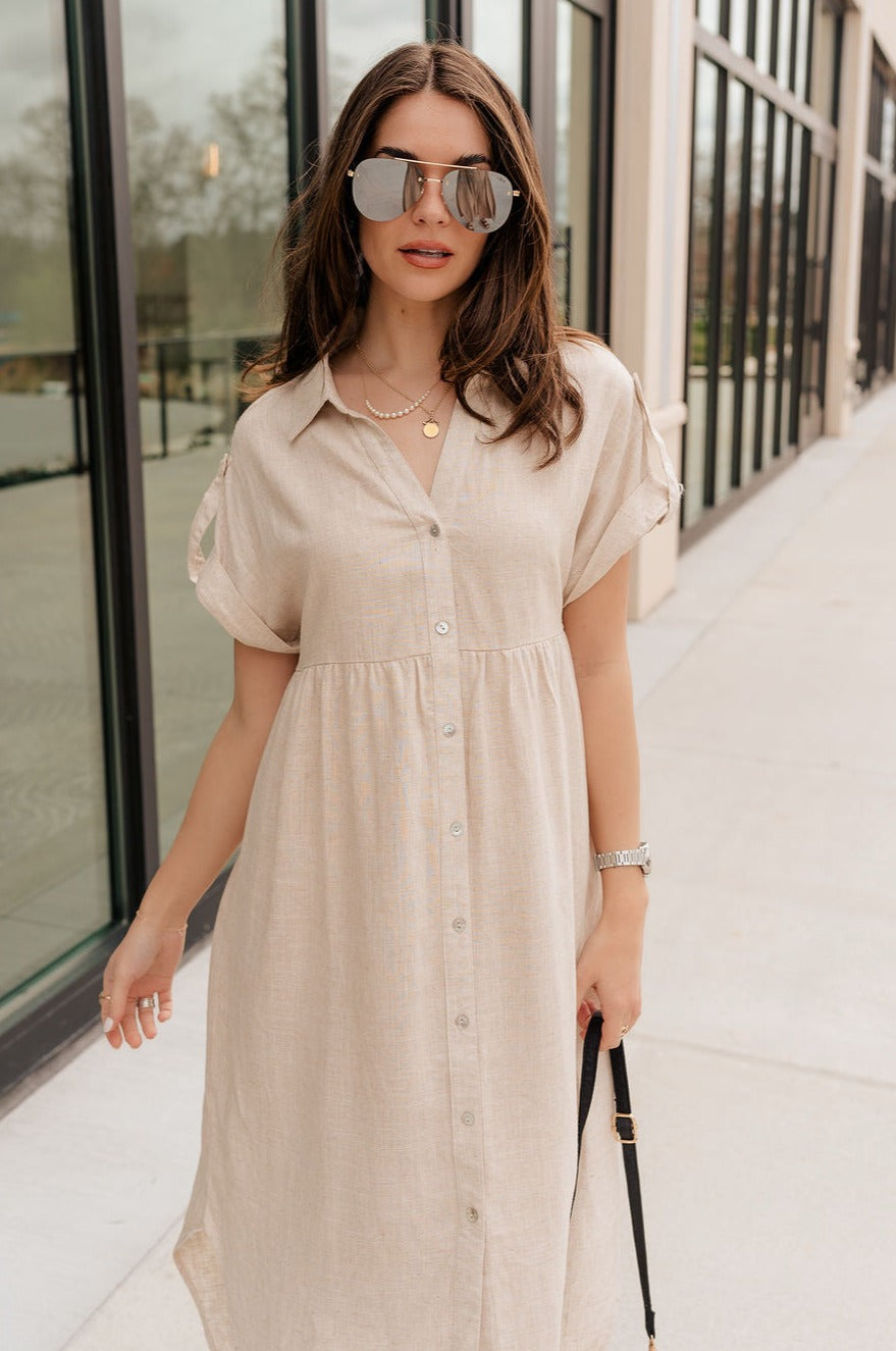 Front view of model wearing the Daphne Oatmeal Button-Up Midi Dress which features beige fabric, a button-up front with pearlescent buttons, a collared neckline, short sleeves with cuffs and button tabs, and a rounded midi hemline.