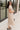 Full body side view of model wearing the Daphne Oatmeal Button-Up Midi Dress which features beige fabric, a button-up front with pearlescent buttons, a collared neckline, short sleeves with cuffs and button tabs, and a rounded midi hemline.